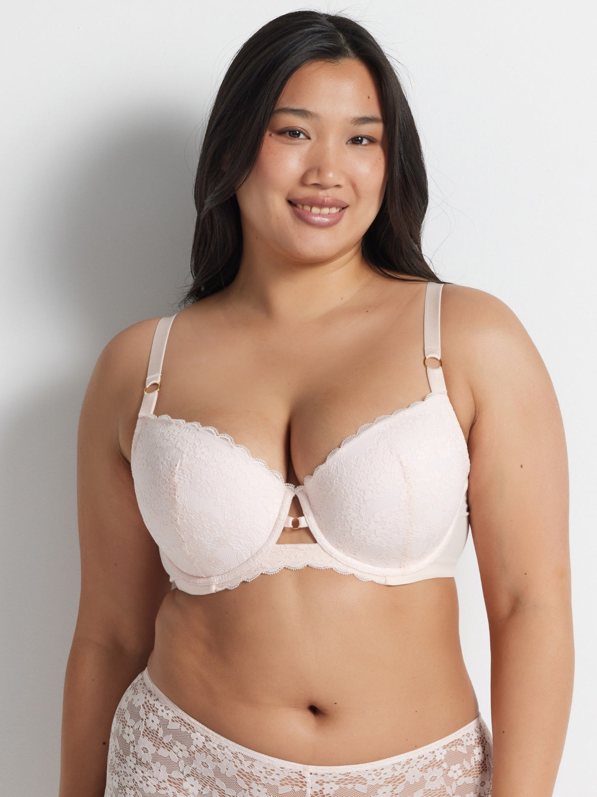 Chloe is wearing the Be Free Lace Balconette Bra in Shell with matching briefs, standing against a grey wall.