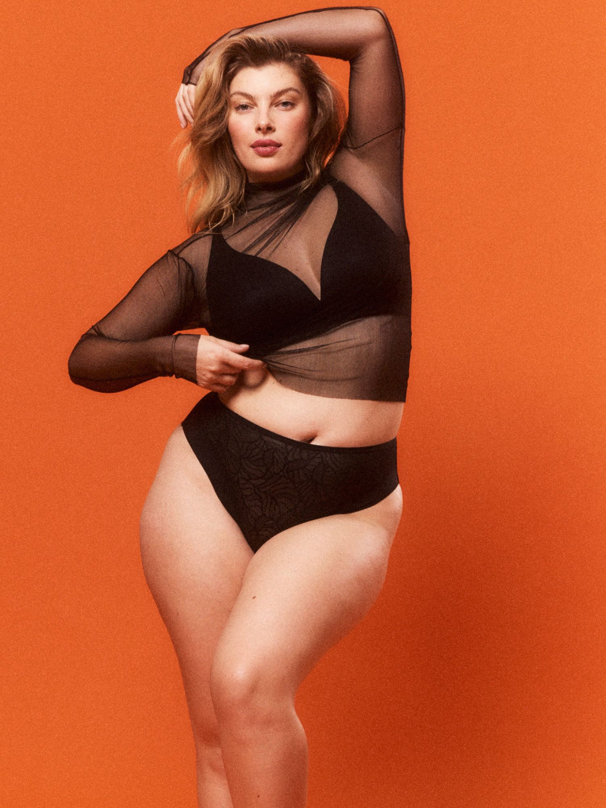 Woman wearing a black sheer top, black wire free bra and high-waisted underwear against an orange background