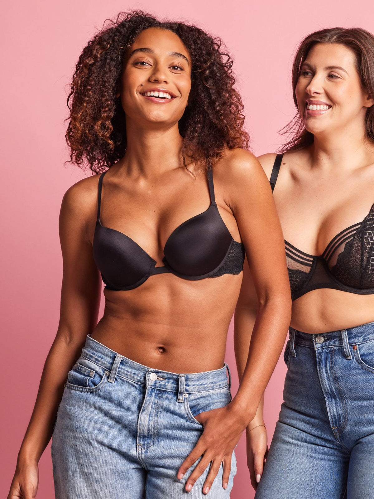 Two women wearing black bras and blue jeans against a pink background